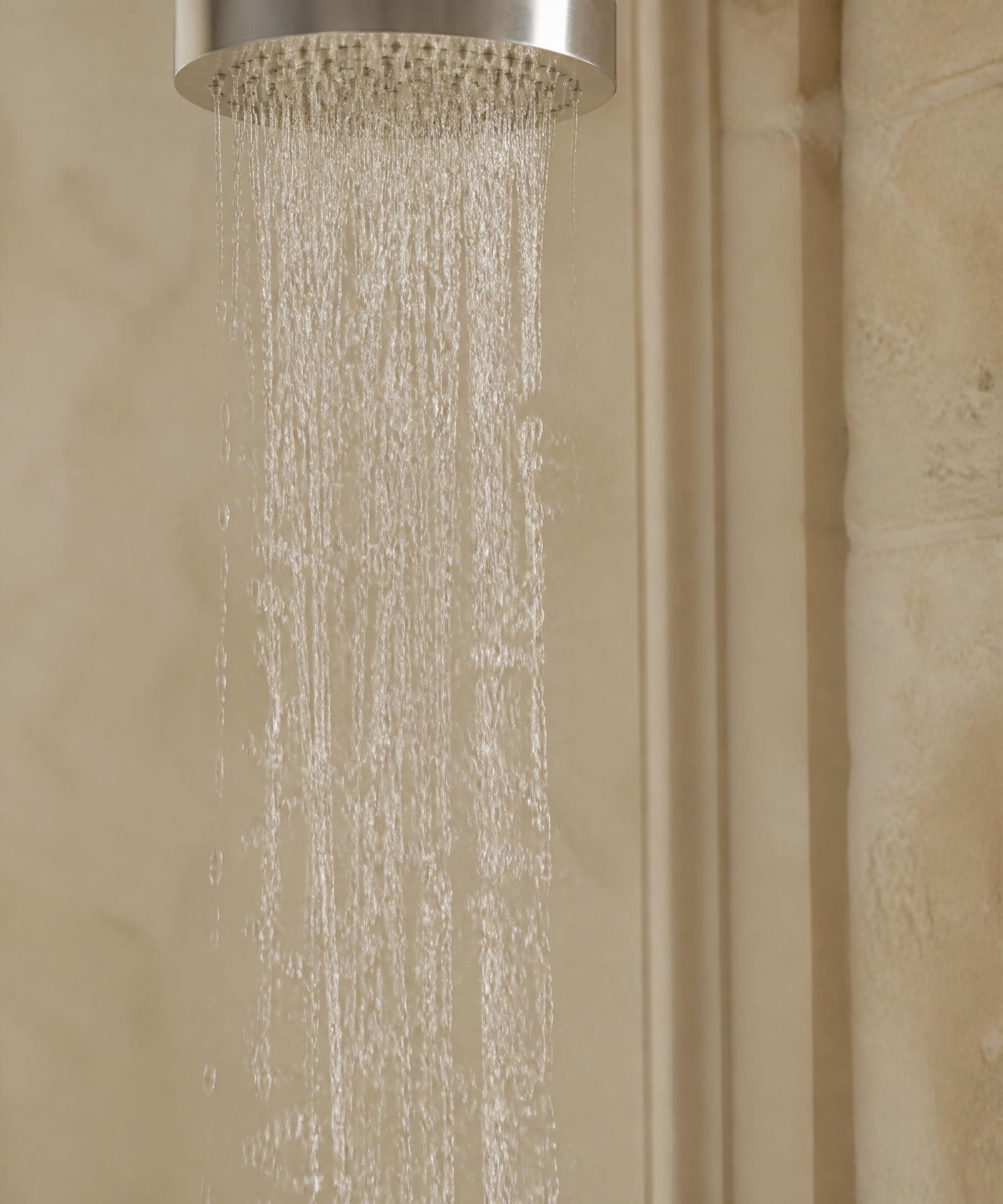Filet d'eau filtrée, limpide et soyeux, tombant d'une pomme de douche en acier brossé devant un mur de pierre calcaire dans un appartement parisien.